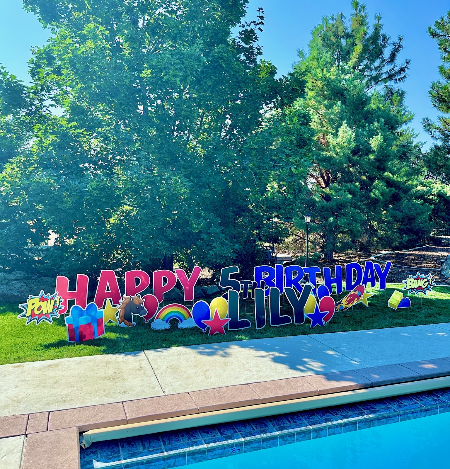 Happy 5th Birthday Lily yard card display with colorful superhero letters and party art beside a Boise Idaho backyard pool